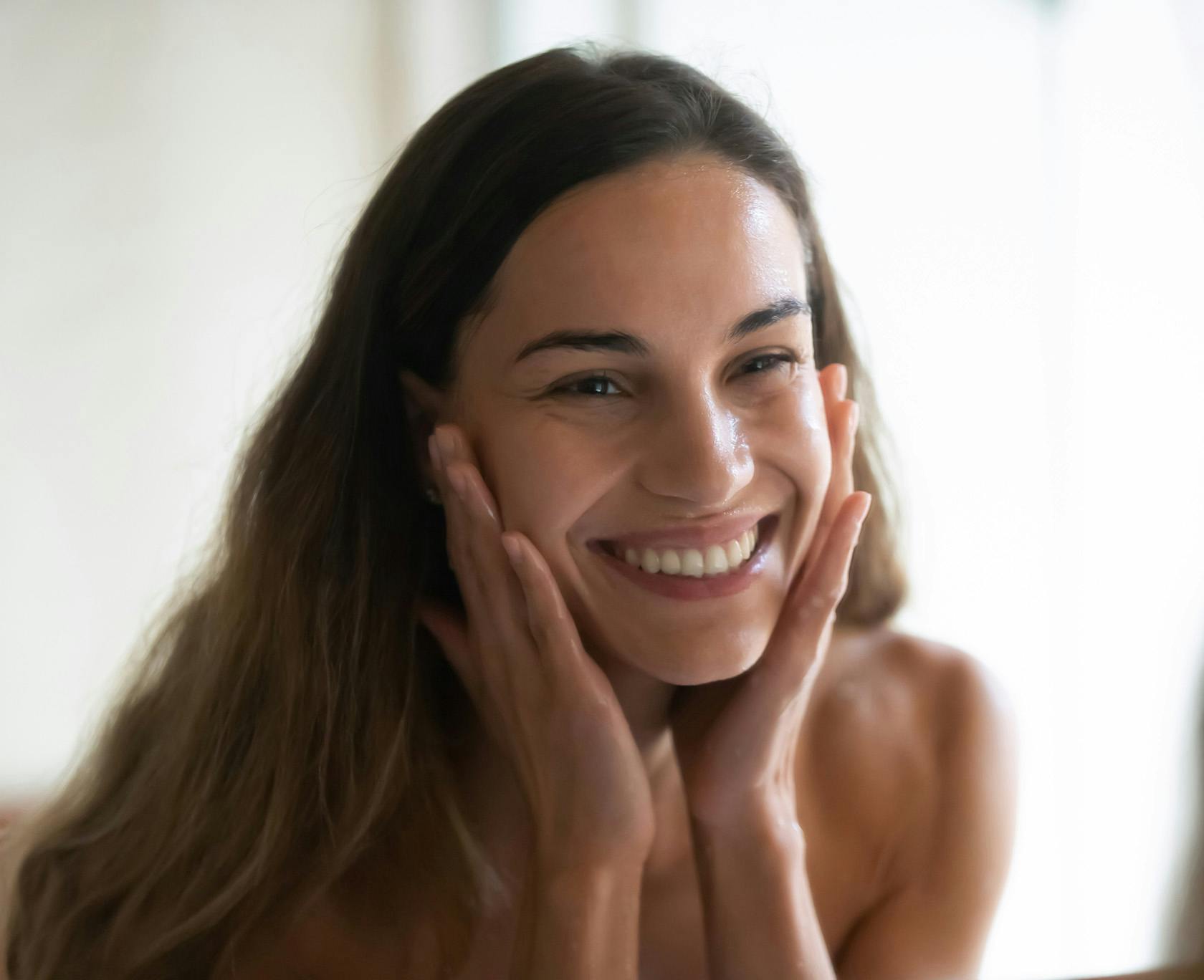 smiling woman with long hair and a white shirt on looking at her reflection in a mirror