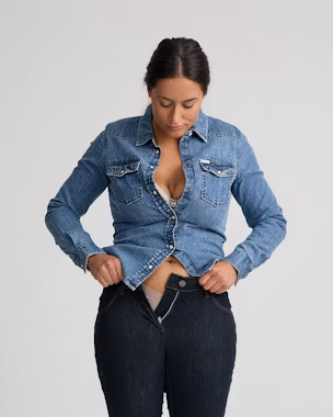 Kleine und kurvige Frau, die eine Jeansbluse und eine High-Waist-Jeans zurechtrückt und damit die Suche nach figurgerechter Kleidung veranschaulicht.