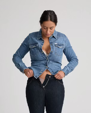Kleine und kurvige Frau, die eine Jeansbluse und eine High-Waist-Jeans zurechtrückt und damit die Suche nach figurgerechter Kleidung veranschaulicht.