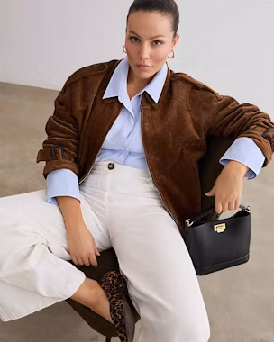 Woman seated in a brown corduroy blouson jacket, light blue shirt and wide white trousers, ballerinas and a black handbag.