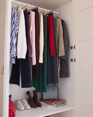 Armadio bianco aperto con camicetta a righe blu e bianche, jeans blu scuro, maglia rosa a maniche lunghe, trench beige e altri capi appesi, sneakers bianche, stivaletti marroni e borsa rossa.