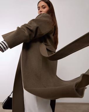 Model trägt einen langen Mantel in Khaki, Gürtel im Wind, Symbol für die Eleganz und fließende Bewegung eines langen Damenmantels.