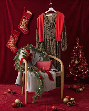 Abito fantasia su una gruccia, maglione rosso, cappotto di lana grigio e borsa rossa su una sedia, decorato in stile natalizio e con sfondo rosso