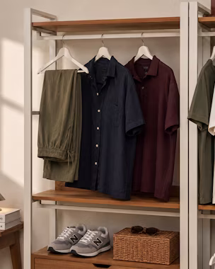 A detailed, warm-lit photograph of a modular wooden and metal wardrobe unit, displaying curated men's outfits in cohesive colors like olive green, navy blue, deep red, and white, demonstrating a complete capsule collection with sneakers and accessories for effortless style coordination.
