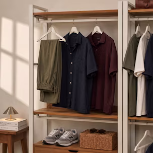 A detailed, warm-lit photograph of a modular wooden and metal wardrobe unit, displaying curated men's outfits in cohesive colors like olive green, navy blue, deep red, and white, demonstrating a complete capsule collection with sneakers and accessories for effortless style coordination.