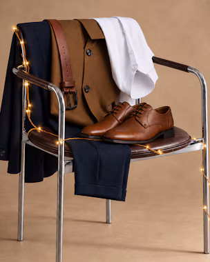Men’s festive outfit with navy suit, white shirt, brown belt and leather shoes arranged on a chair.