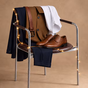 Look festivo de hombre con traje azul marino, camisa blanca, cinturón marrón y zapatos de piel sobre una silla.
