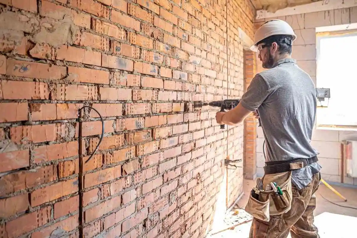 Un artisan sur un chantier de rénovation utilise une carotteuse sur un mur en briques pour créer un déficit foncier.