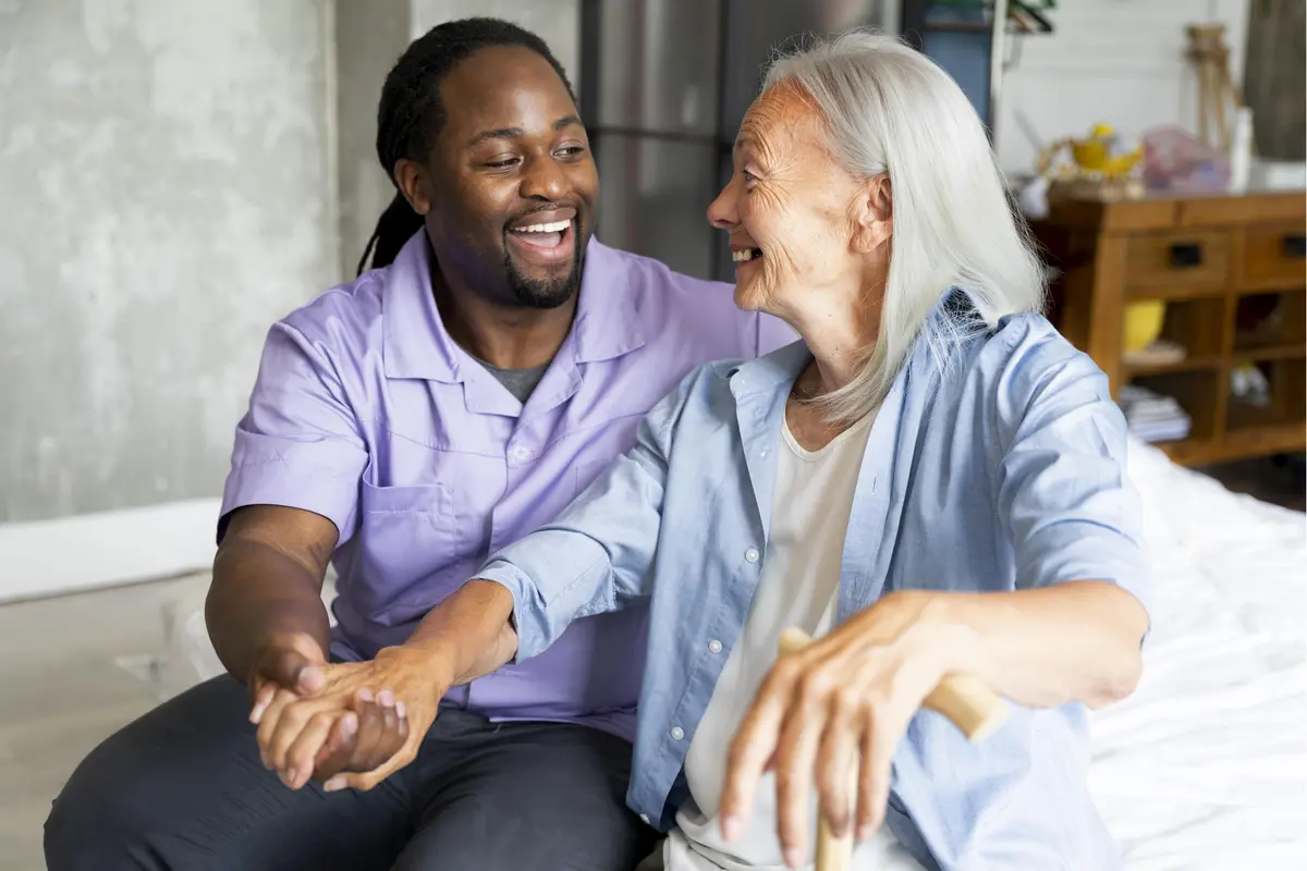 Aide-soignant souriant en blouse violette tenant la main d'une femme âgée avec une canne, illustrant l'accompagnement humain pour un investir en résidence EHPAD.