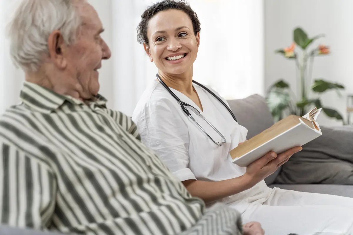 Infirmière souriante avec un stéthoscope tenant un livre ouvert et échangeant avec un homme âgé, illustrant les services de soins pour investir en résidence EHPAD.
