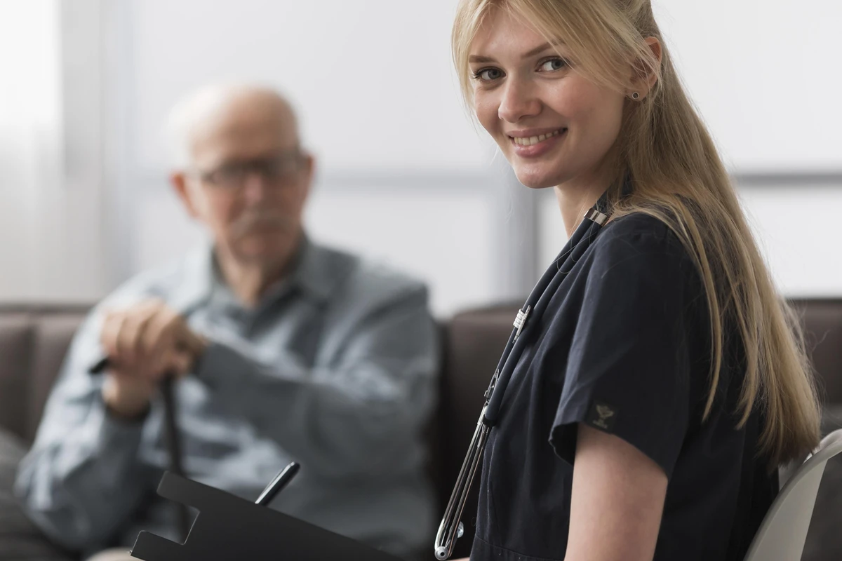 Jeune infirmière blonde souriante avec un stéthoscope au premier plan, avec un homme âgé assis en arrière-plan, idéal pour illustrer un projet pour investir en résidence EHPAD.