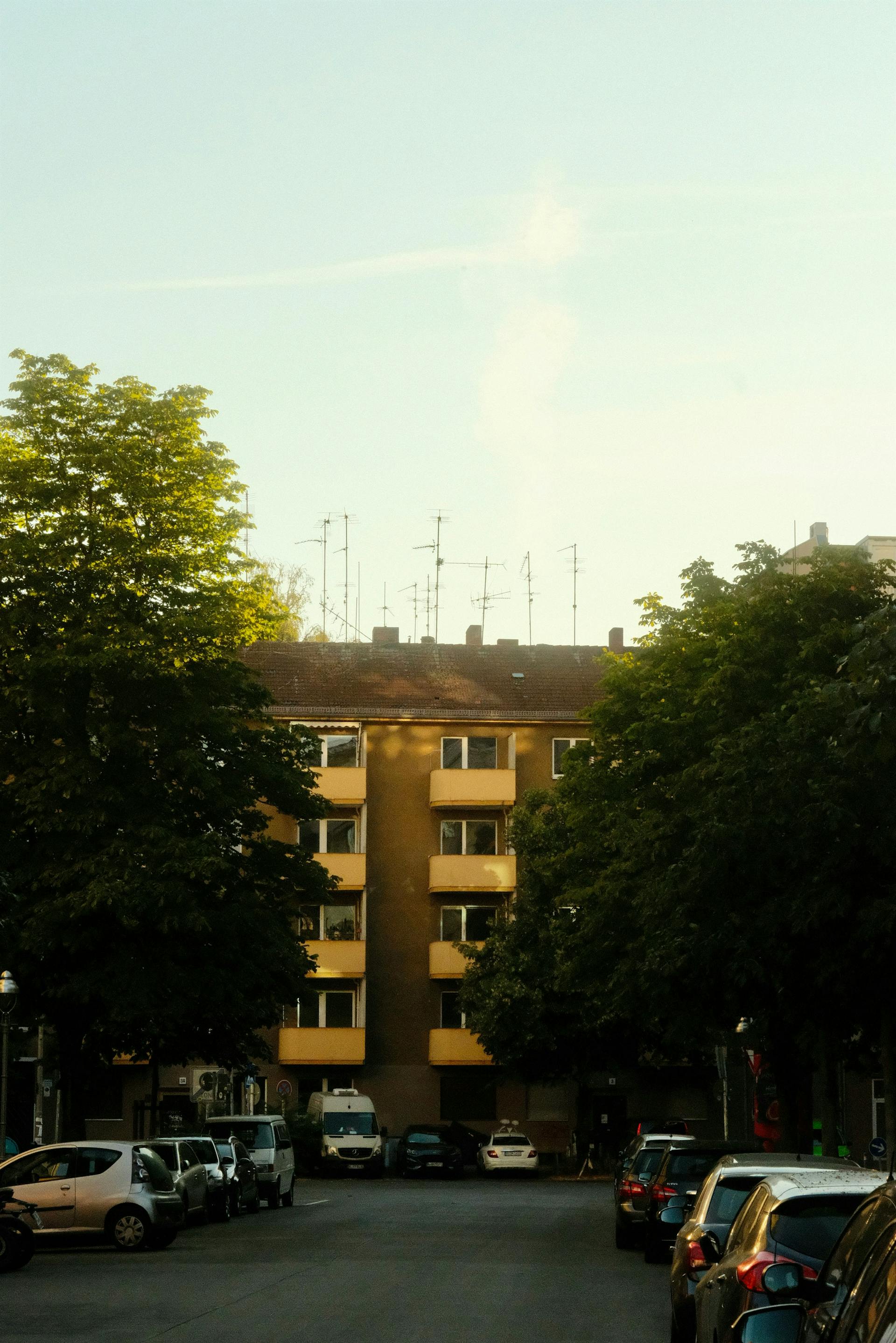 Residential street with parked cars and trees in front of a yellow apartment building with balconies in evening light.