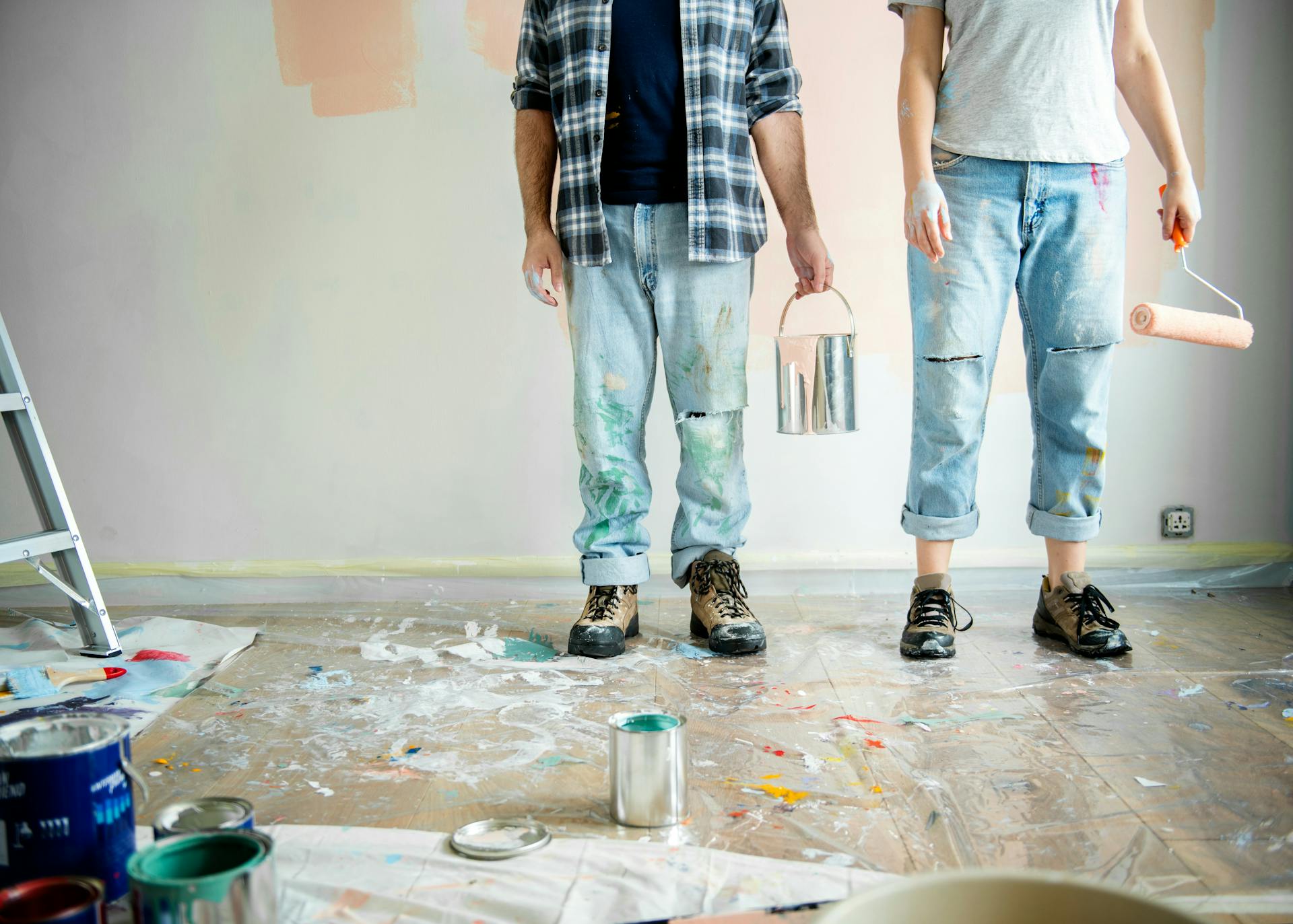 Couple painting a wall together in an empty apartment, holding paint cans and a roller.