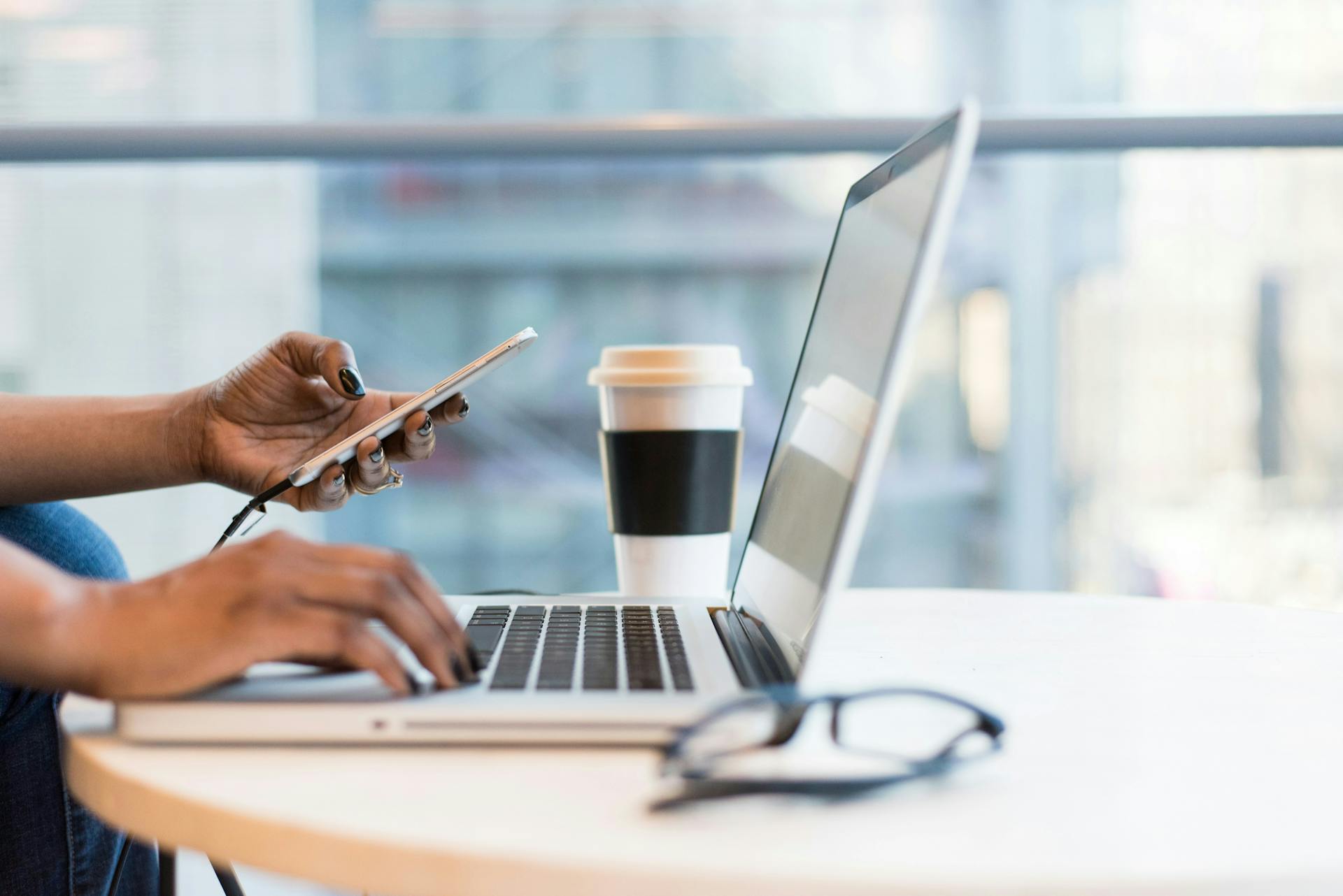 Person tippt auf einem Laptop und hält gleichzeitig ein Smartphone in der Hand, daneben ein Kaffeebecher.