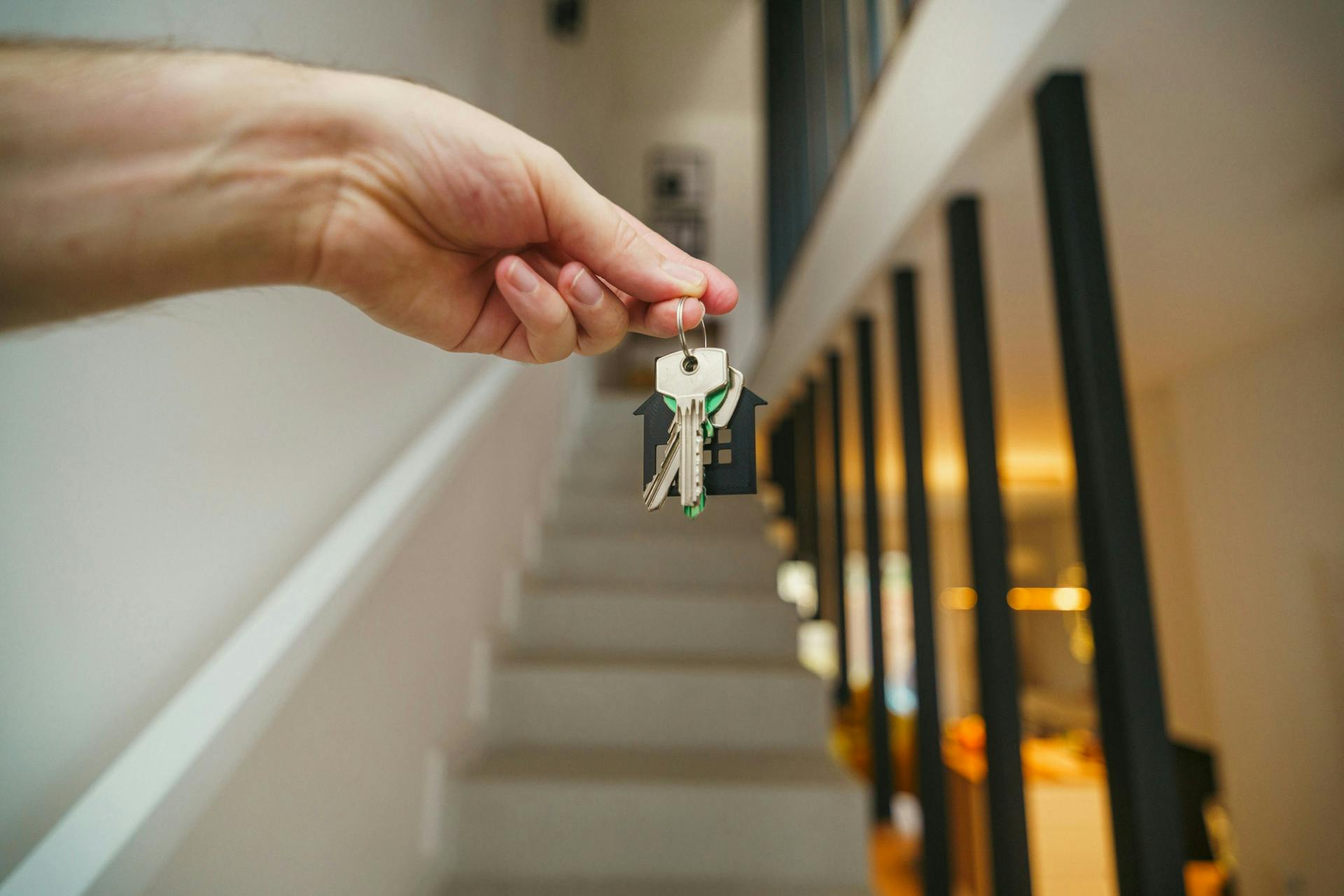A hand holds an apartment key with a house-shaped keychain in a stairwell.