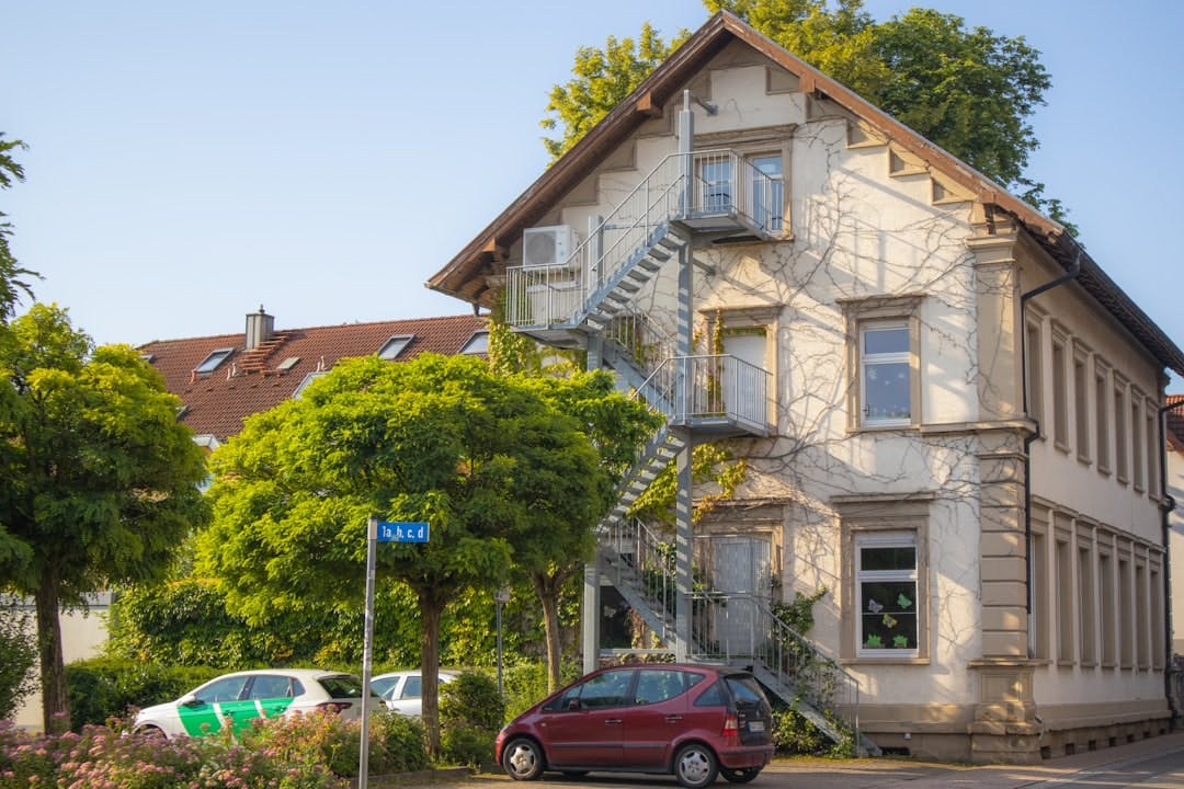 Cremefarbenes Altbauhaus mit außenliegender Metalltreppe und Efeu an der Fassade, Bäume und parkende Autos davor.