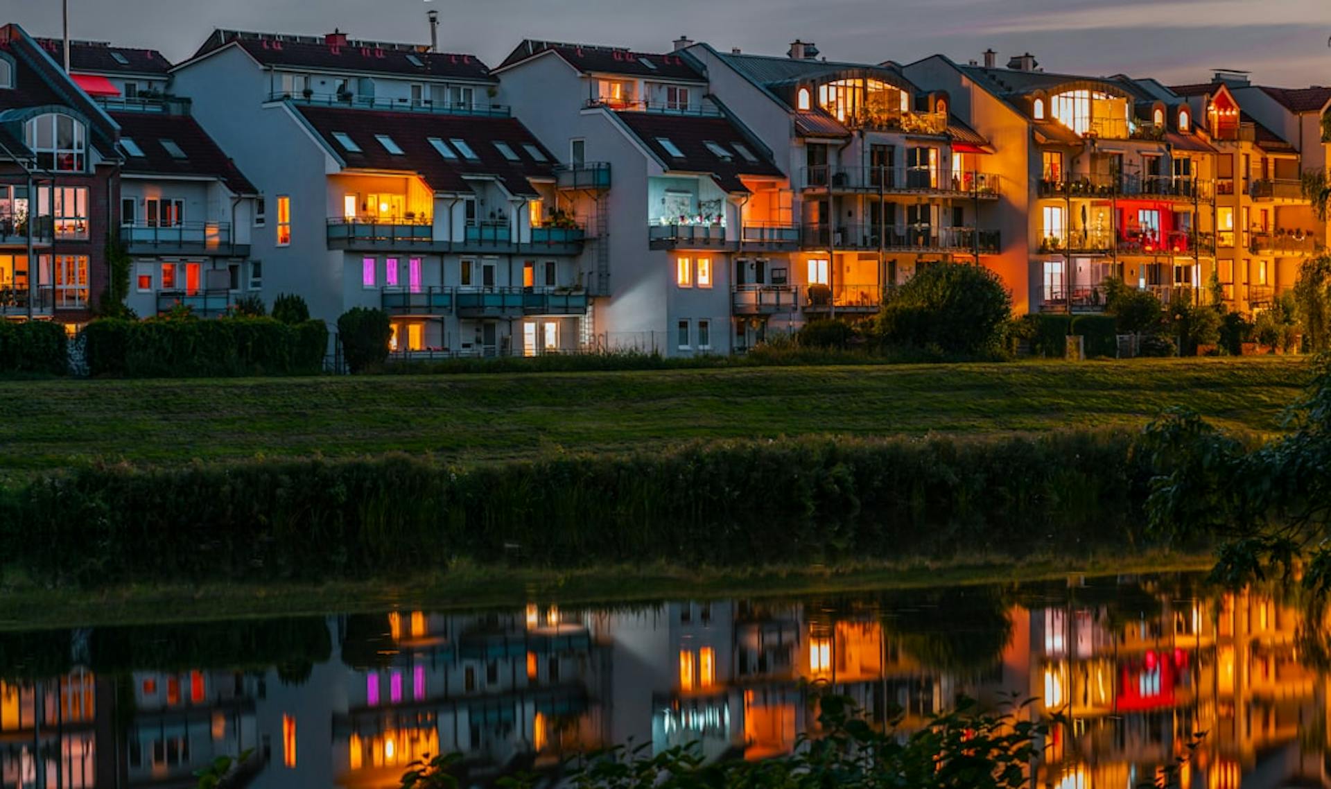 Illuminated apartment buildings by the water at dusk, reflected in the surface.