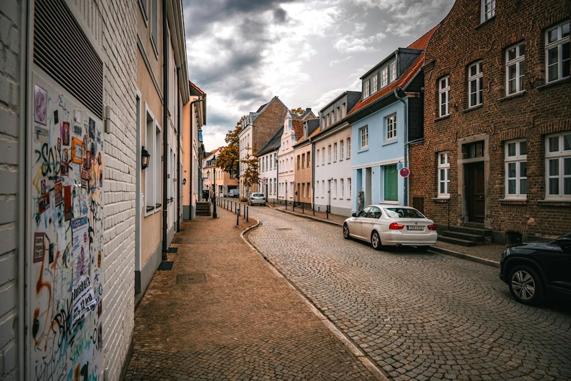 Cobblestone street lined with colorful houses under a cloudy sky, cars parked along the curb.