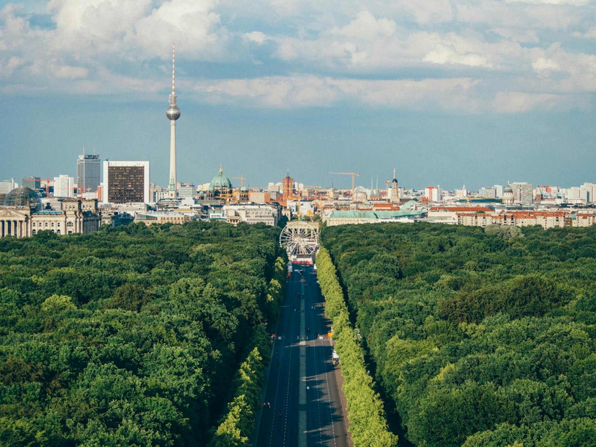 Luftaufnahme des Berliner Tiergartens mit einer Allee, einem Riesenrad und der Skyline mit Fernsehturm im Hintergrund.