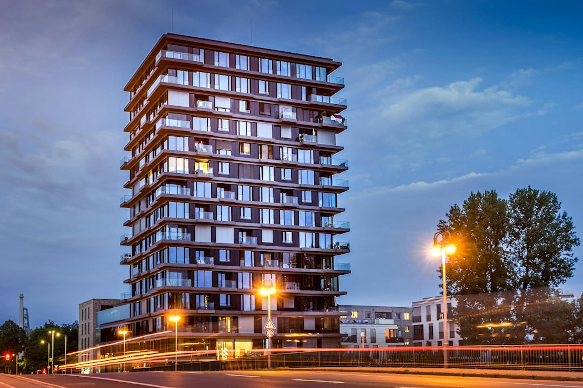 Modernes Hochhaus mit Glasbalkonen bei Dämmerung, im Vordergrund eine Straße mit Lichtspuren.