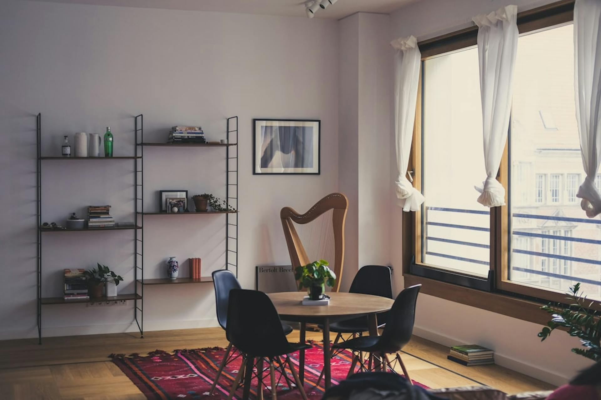 Bright living room with a shelving unit, round dining table, chairs and large curtained windows.