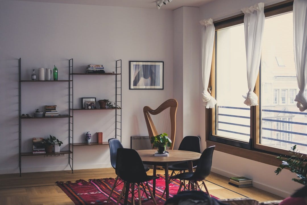 Bright living room with a shelving unit, round dining table, chairs and large curtained windows.
