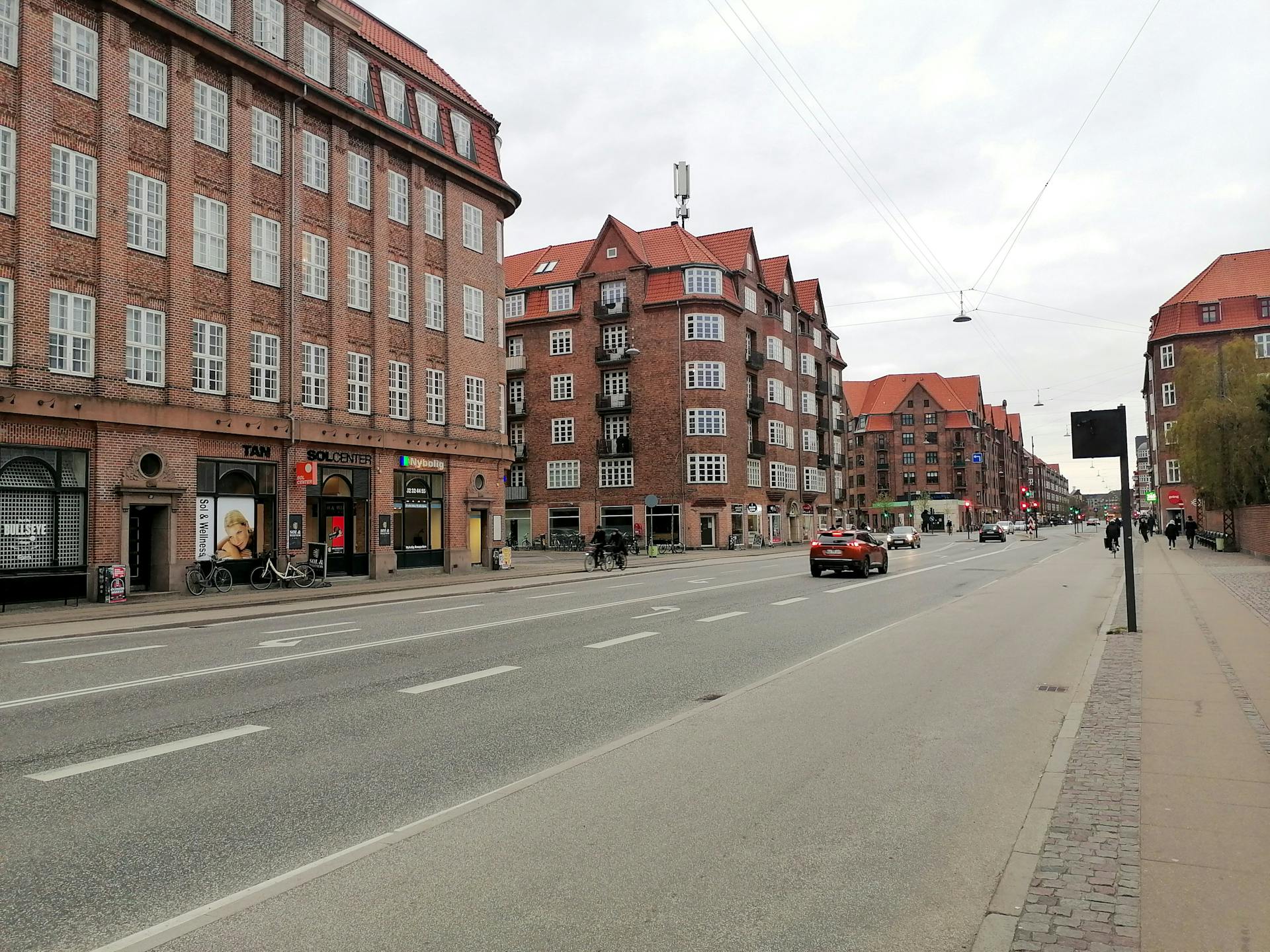 En bred dansk gade med rødstensejendomme, cyklister og en bil, der kører forbi. (Photo via Unsplash)