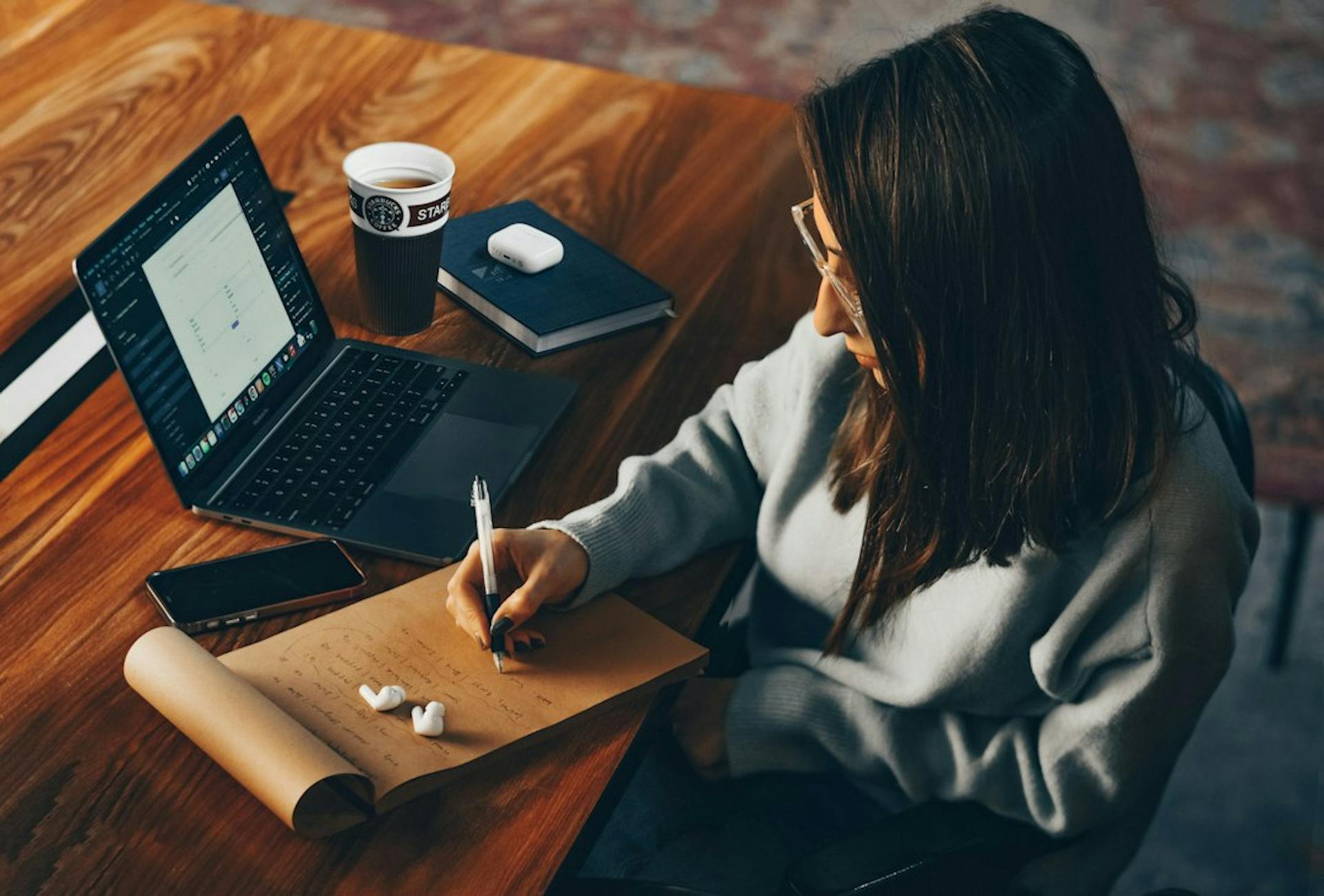 Junge Frau schreibt am Holztisch Notizen neben ihrem Laptop (Photo by Nasim Keshmiri on Unsplash)