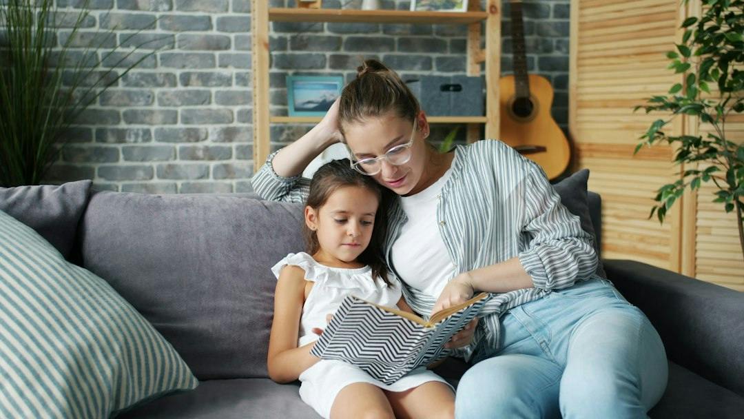 Mother and young daughter reading a book together on the sofa at home (Photo by Vitaly Gariev on Unsplash)