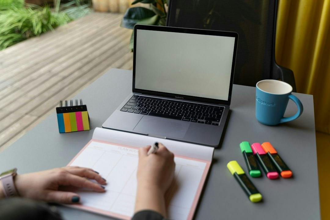Person writing notes in a notebook next to a laptop and coloured markers (Photo by Flipsnack on Unsplash)