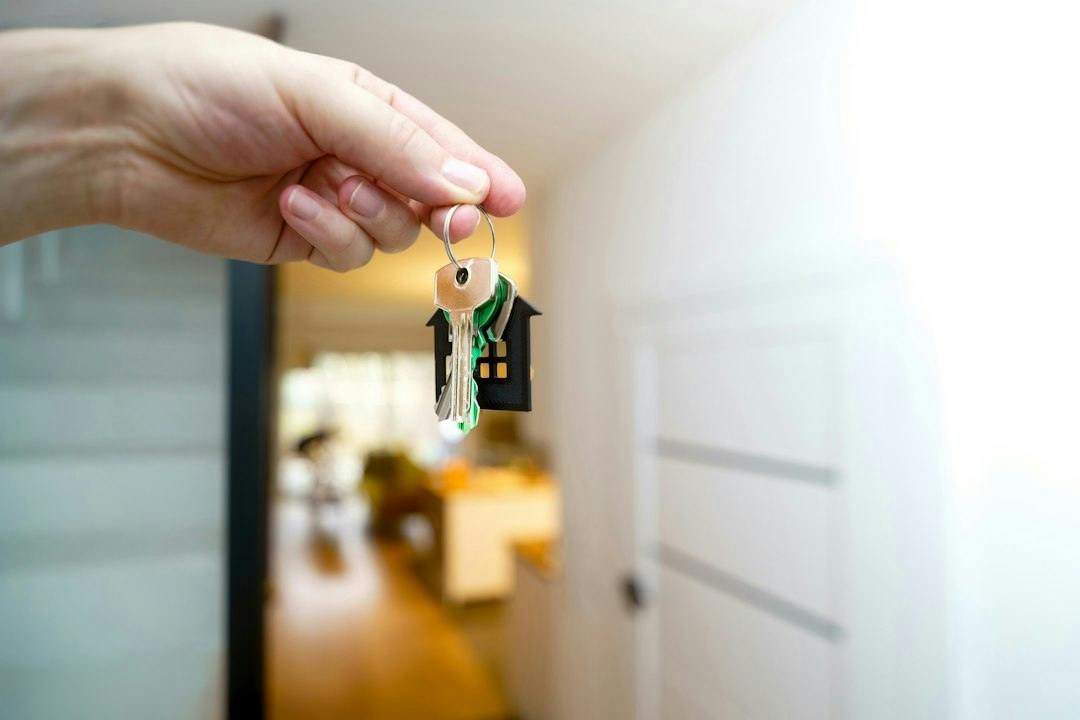 Hand holding house keys with a house-shaped keyring at an apartment entrance (Photo by Jakub Żerdzicki on Unsplash)