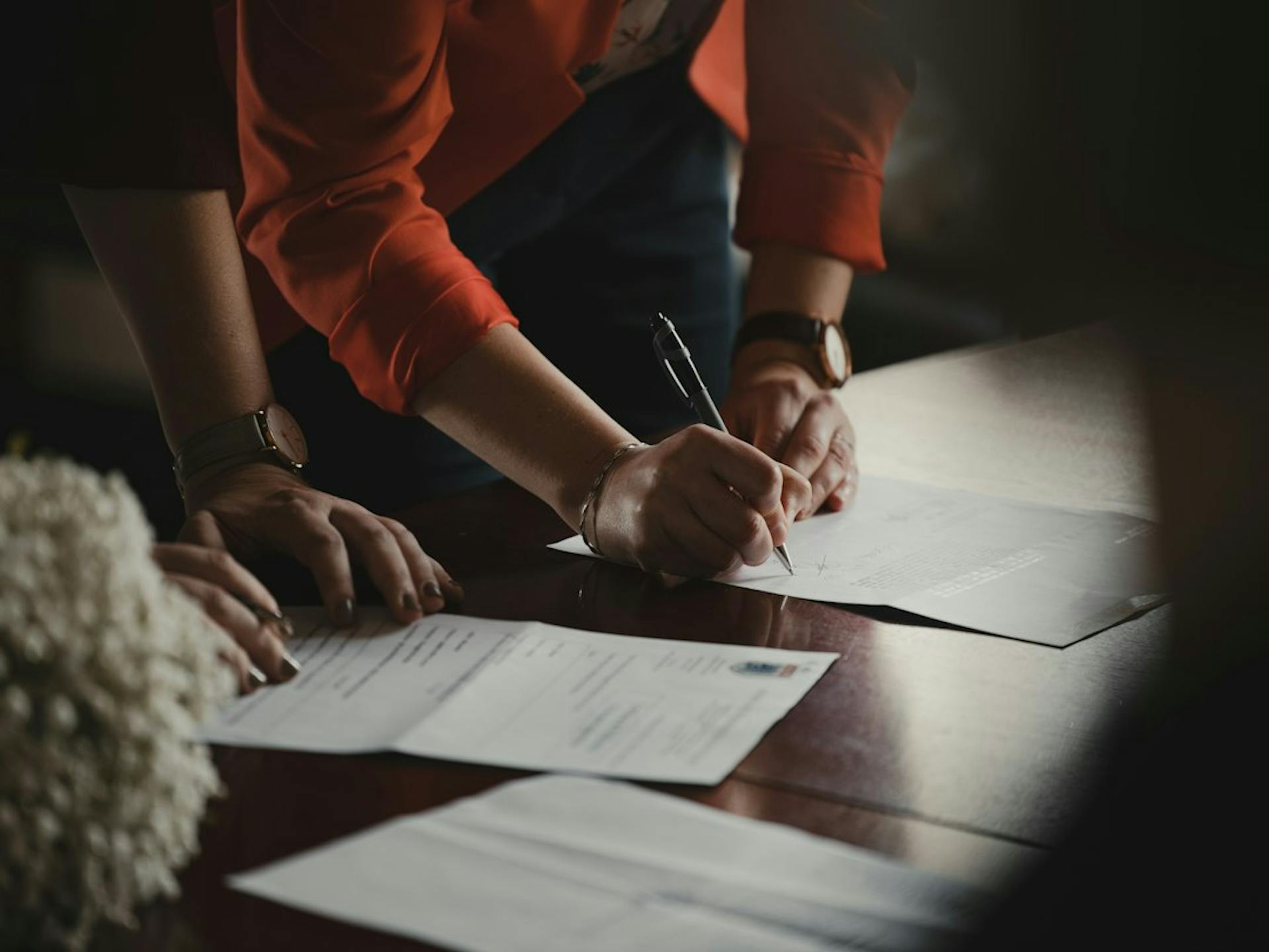 Hands signing paper documents on a dark wooden table (Photo by Romain Dancre on Unsplash)