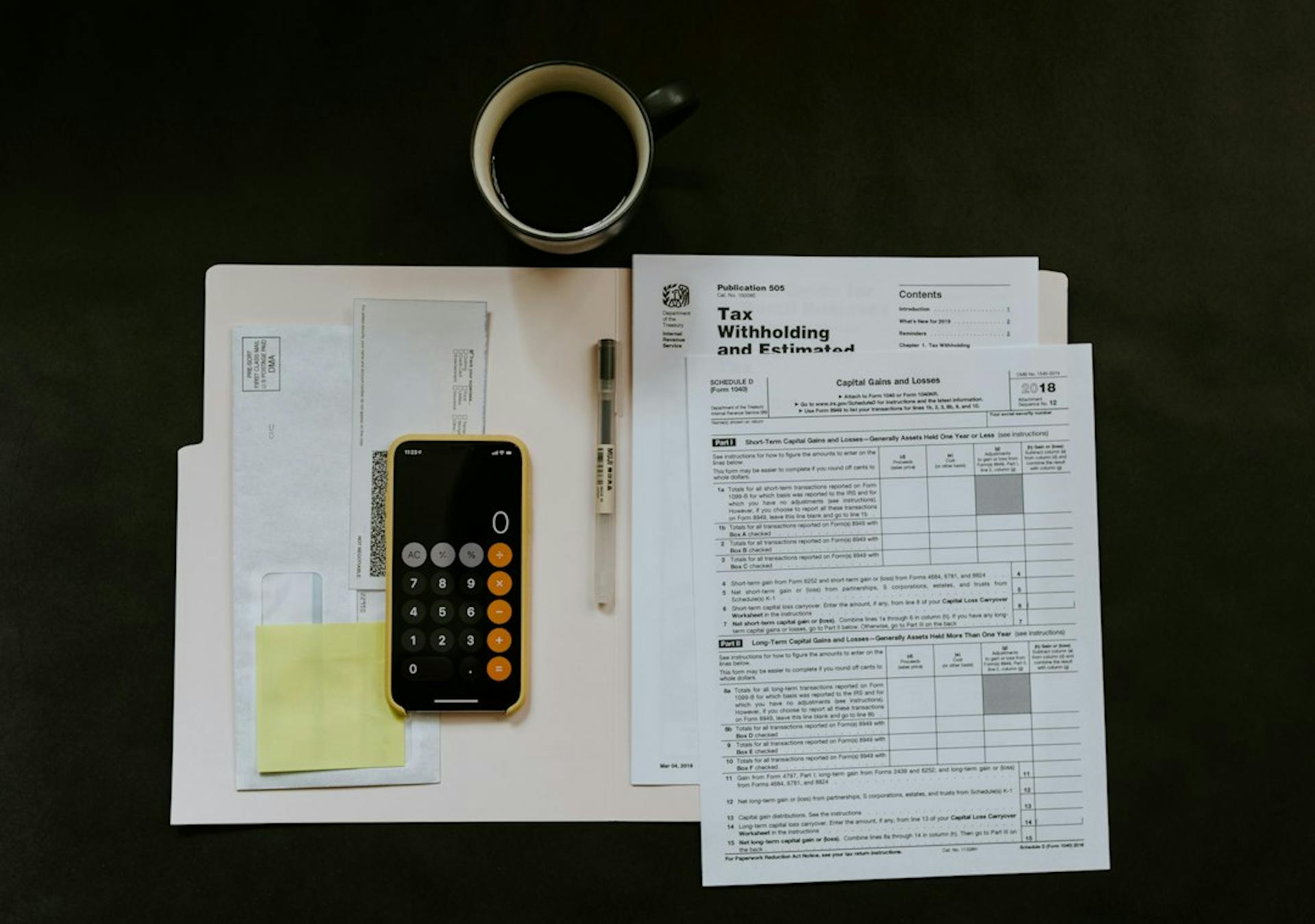 Tax forms, a phone calculator and a coffee cup on a dark table (Photo by Kelly Sikkema on Unsplash)