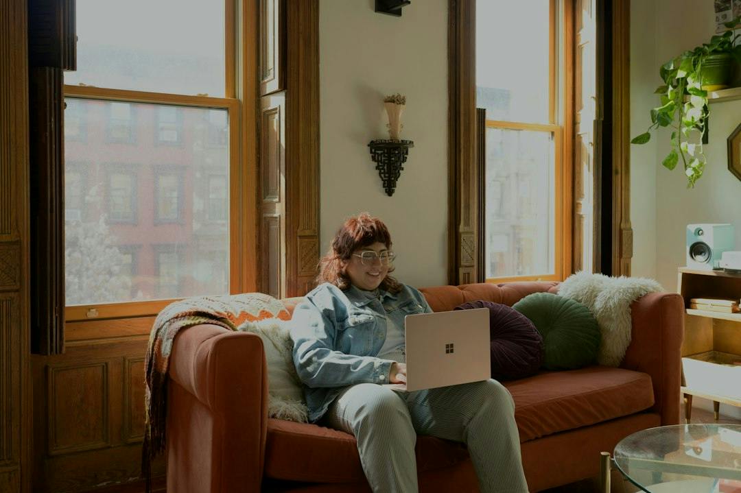 Ung person arbejder på en bærbar i en sofa ved høje vinduer (Photo by Surface on Unsplash)
