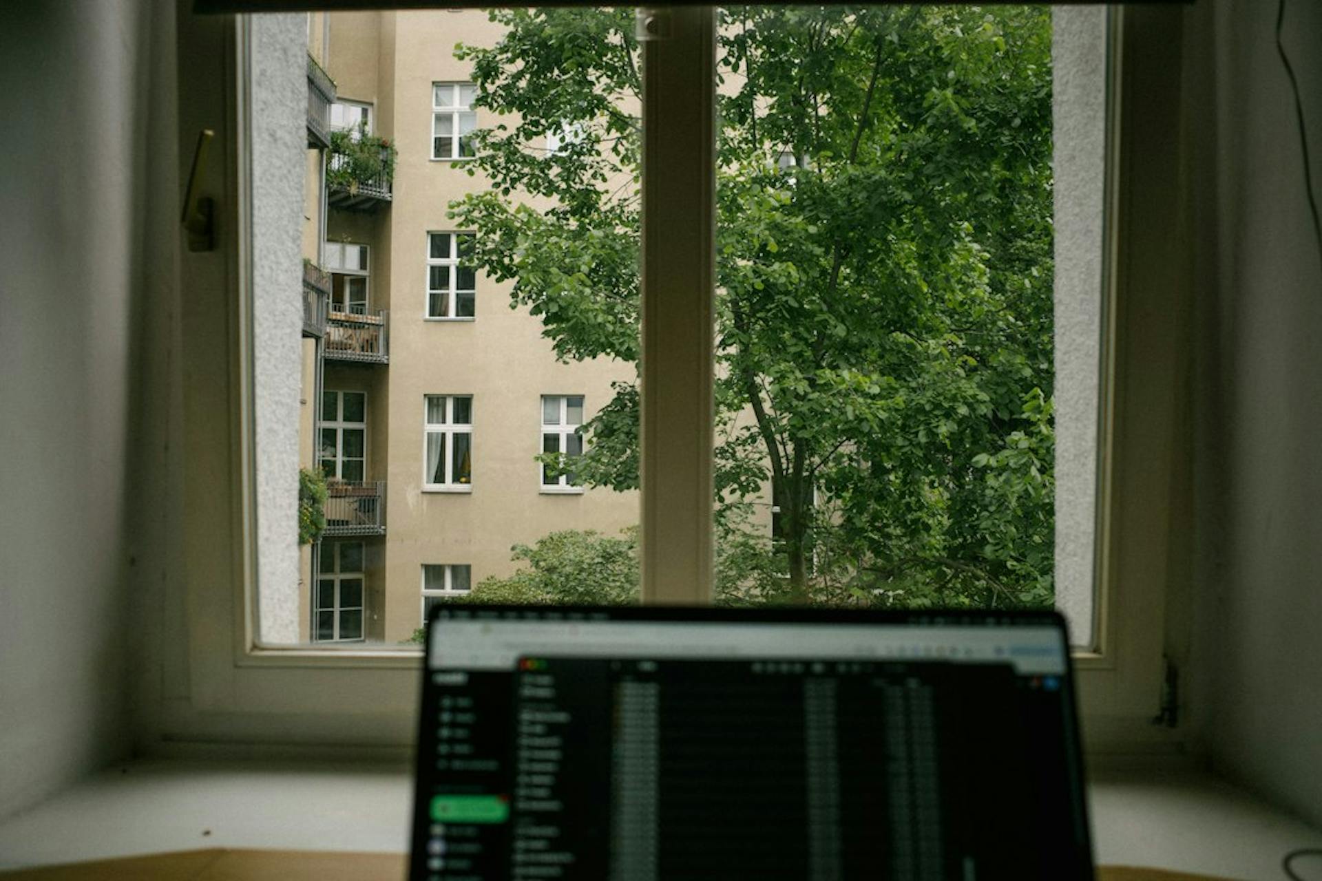 Laptop in front of a window overlooking a leafy apartment courtyard (Photo by Teo Peter on Unsplash)