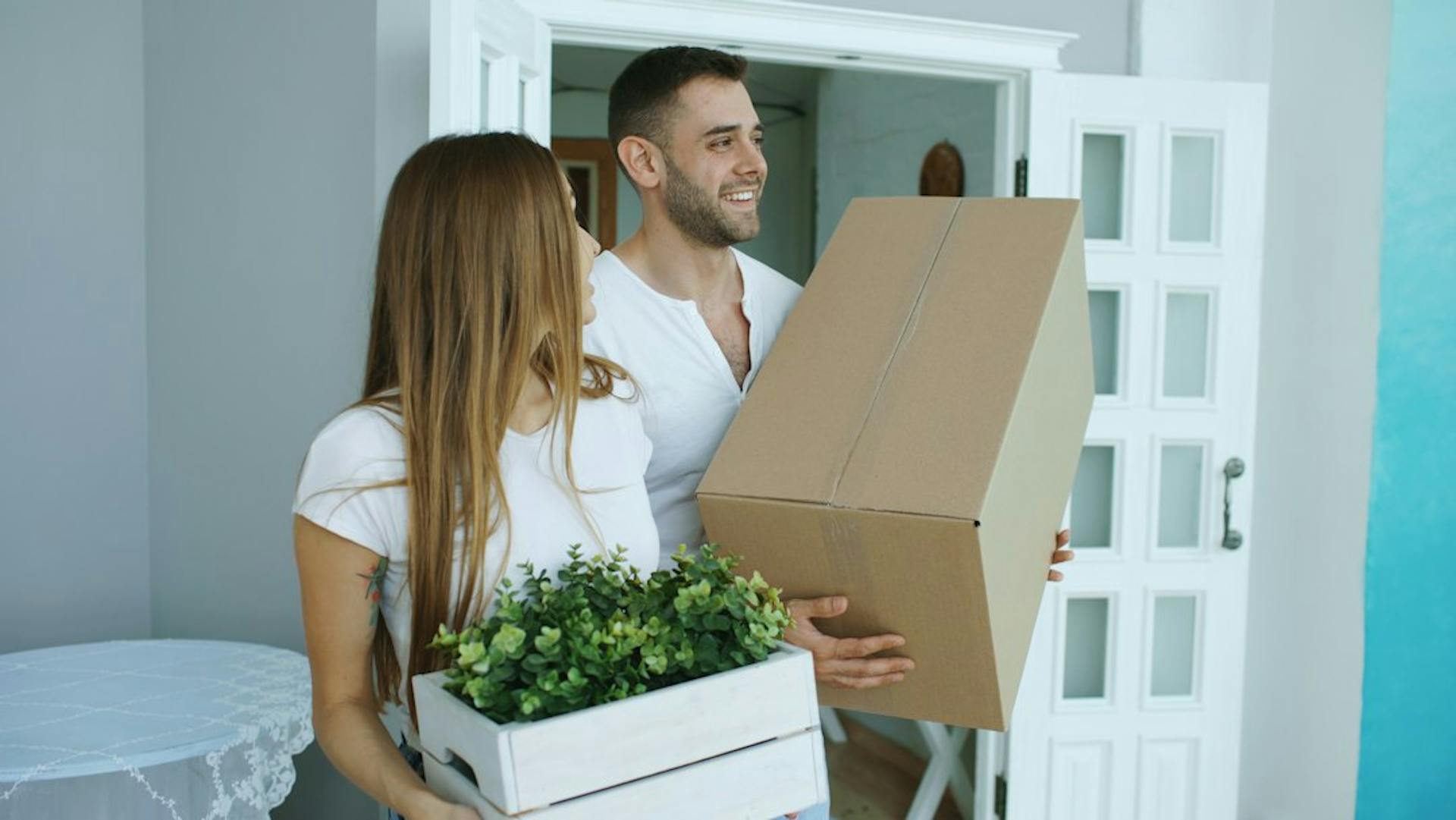 Smiling young couple carrying a moving box and a plant (Photo by Vitaly Gariev on Unsplash)