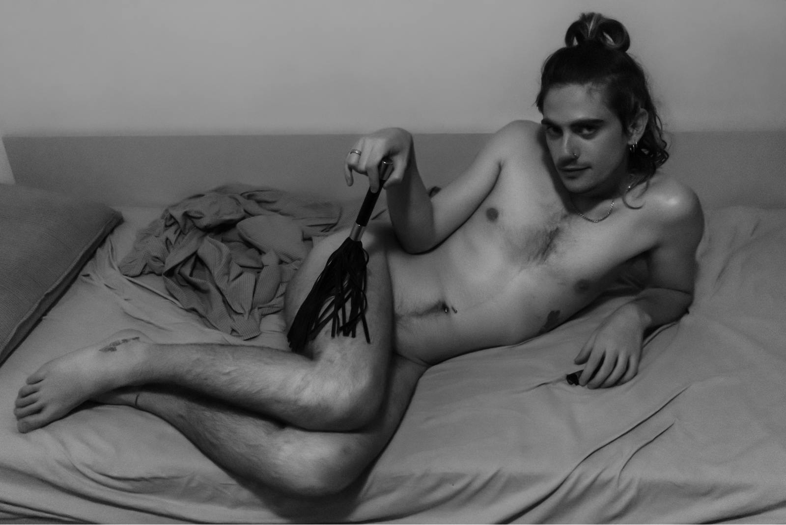 Črno bela fotografija. Gol mlad moški na boku leži na postelji in se opira na komolec. V drugi roki drži bič, v kamero gleda izzivalno.