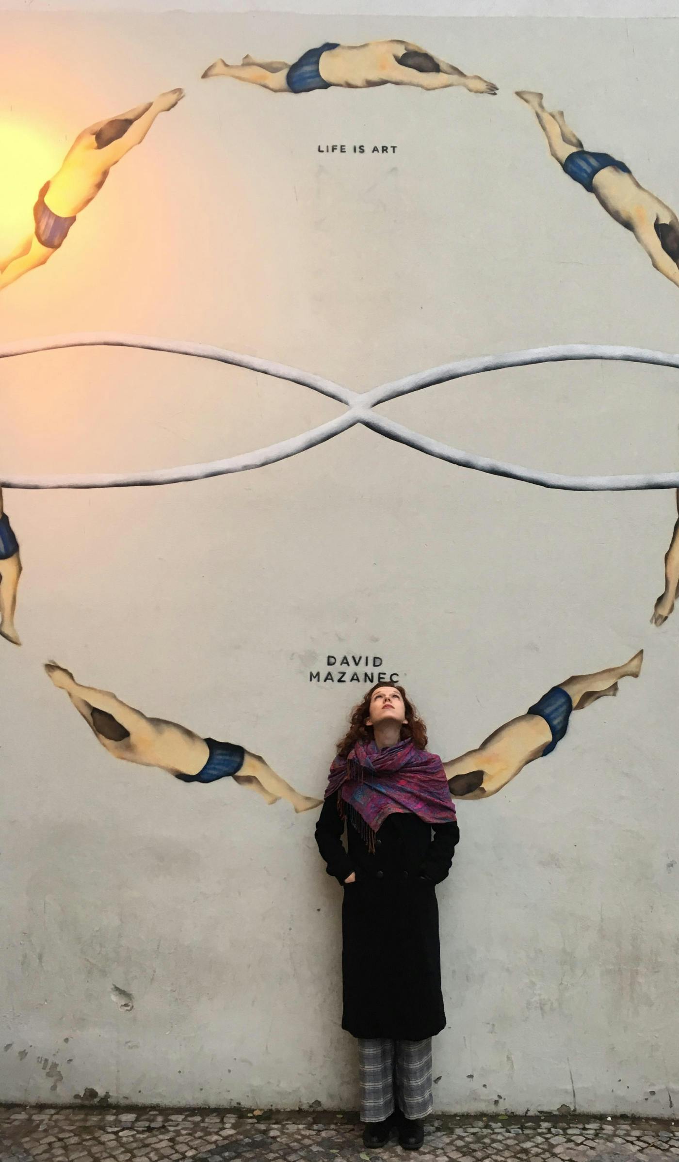 Mlada ženska stoji pred steno z grafiti plavalcev v krogu, za njeno glavo podpis avtorja David Mazanec, nad njo pa napis Life is art
