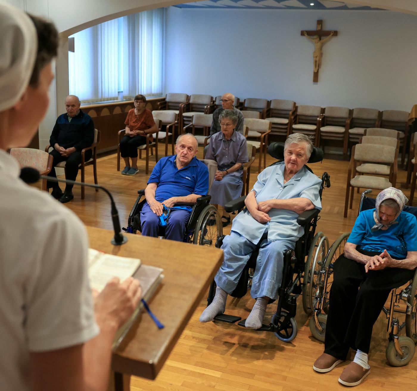 Redovnica bere iz knjige skupini starejših ljudi, trije od njih so na invalidskem vozičku