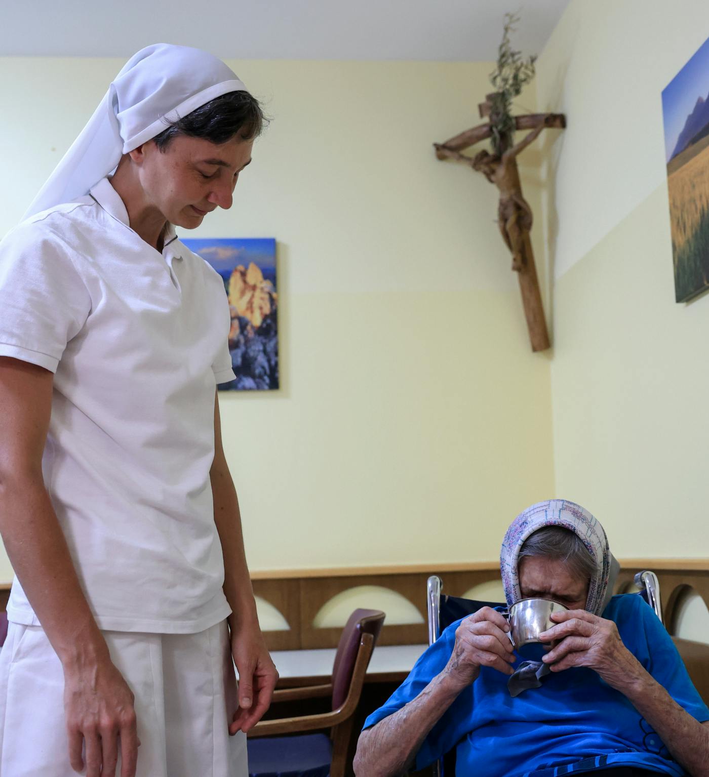 Redovnica v belih oblačilih stoji pred starejšo žensko, ki sedi in pije iz skodelice