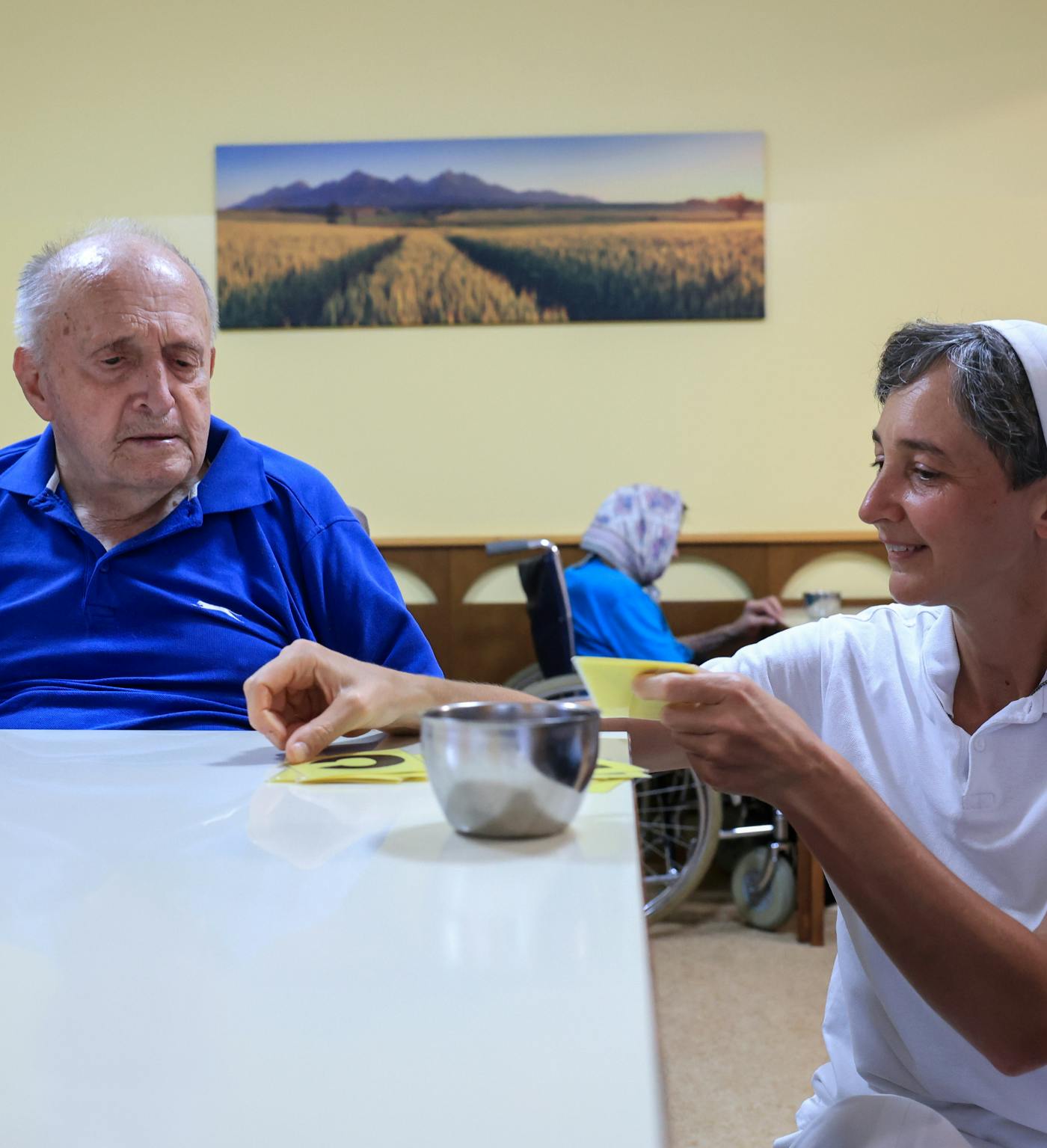 Redovnica čepi pred mizo, za katero sedi starejši moški, pred njega polaga kartice s številkami