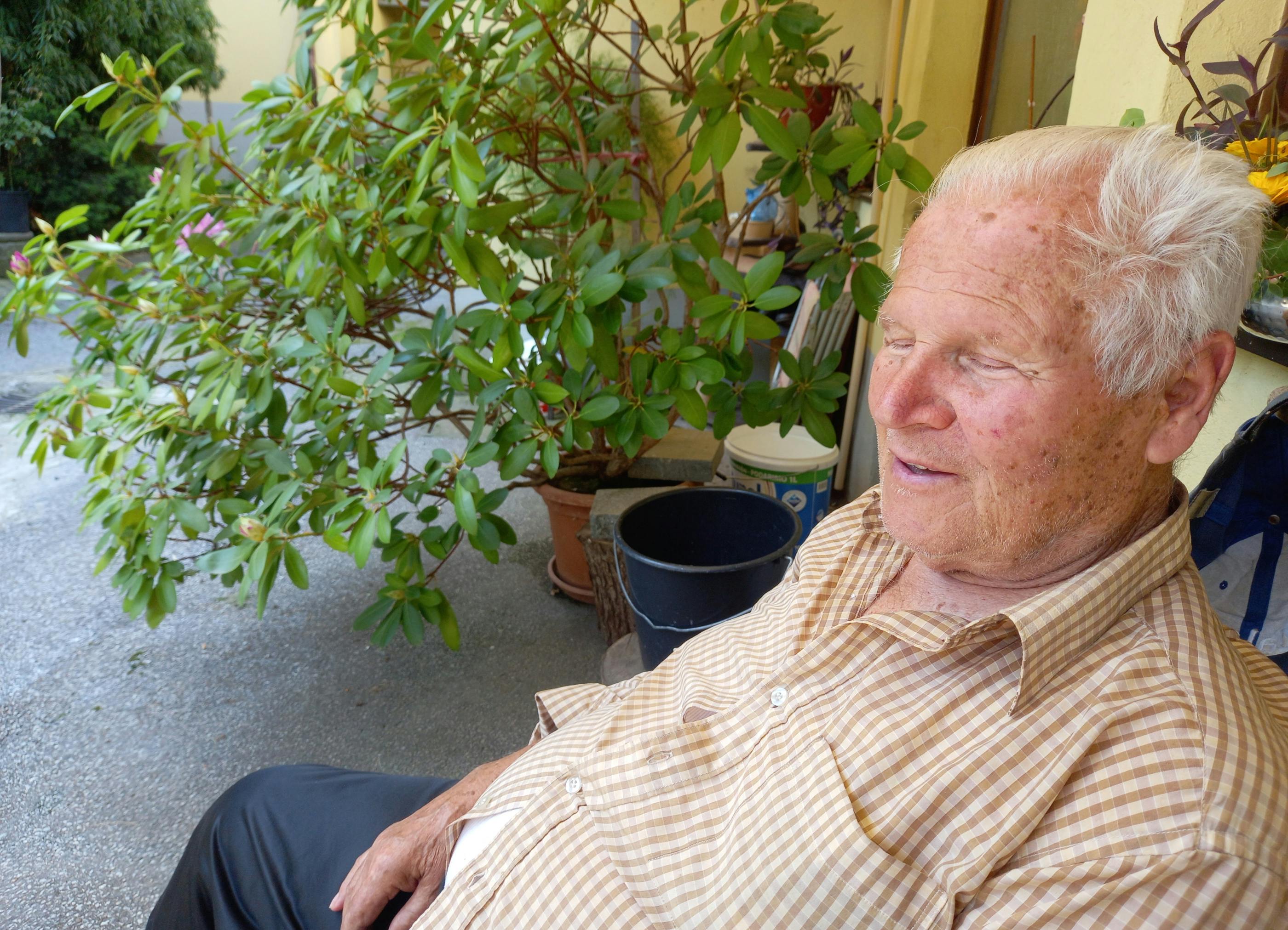 Starejši gospod, ki sedi na terasi za hišo, za njim manjše drevo