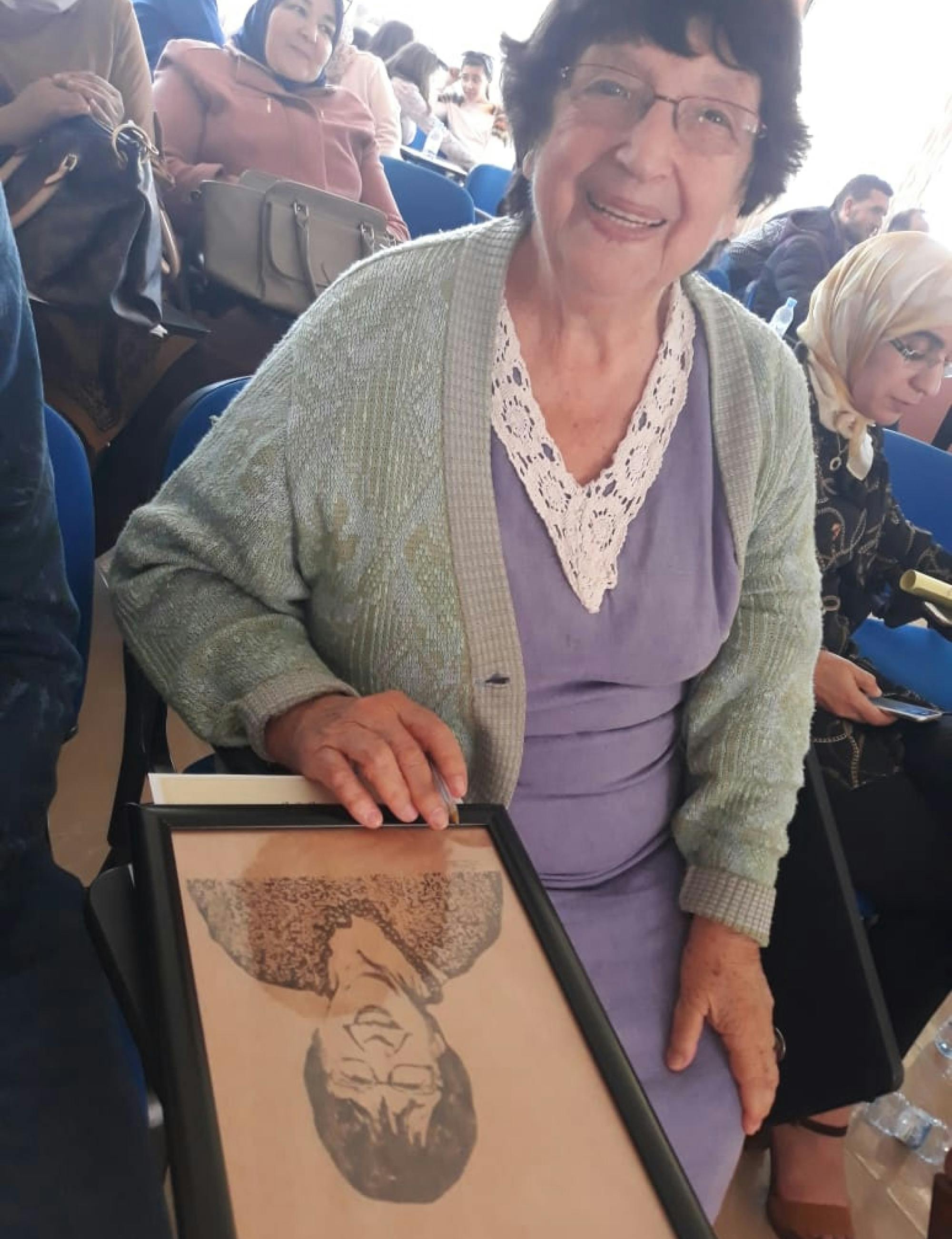 dr. Mikuš Kos, sedi in pred seboj ima uokvirjeno ilustracijo sebe. Nasmejano gleda v kamero