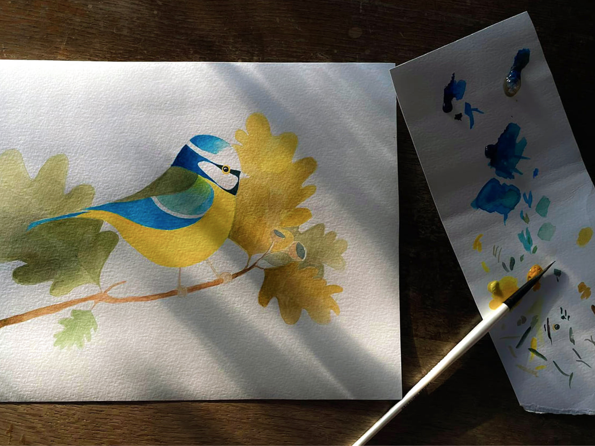 Ilustracija ptiča na belem papirju. Zraven čopič in papir z različnimi odtenki barv.