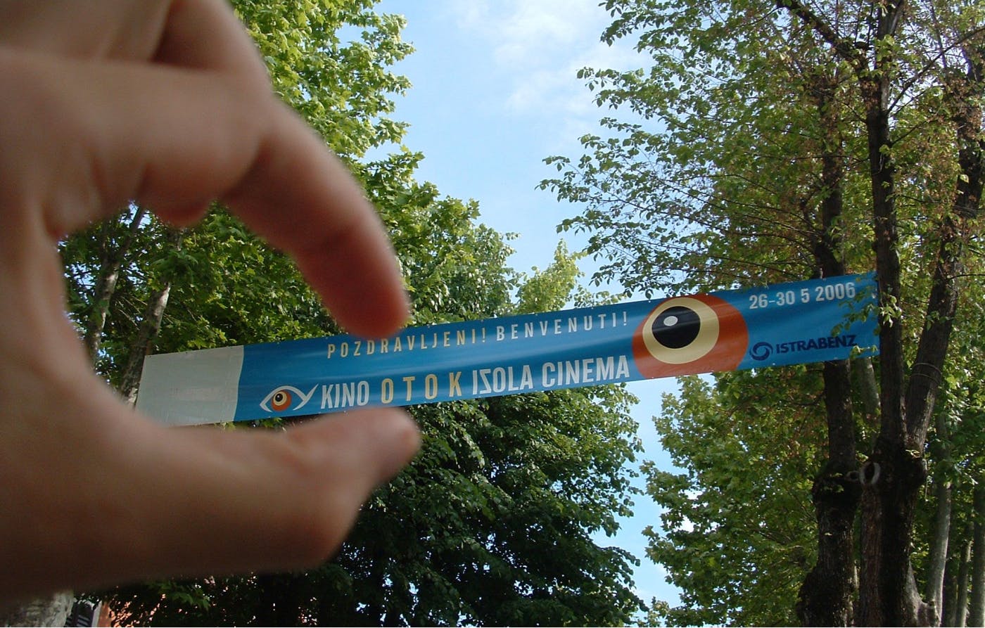 Razpet plakat Kino Otoka 2006 nad ulico, avtor ustvarja iluzijo, kot da ga drži s prsti