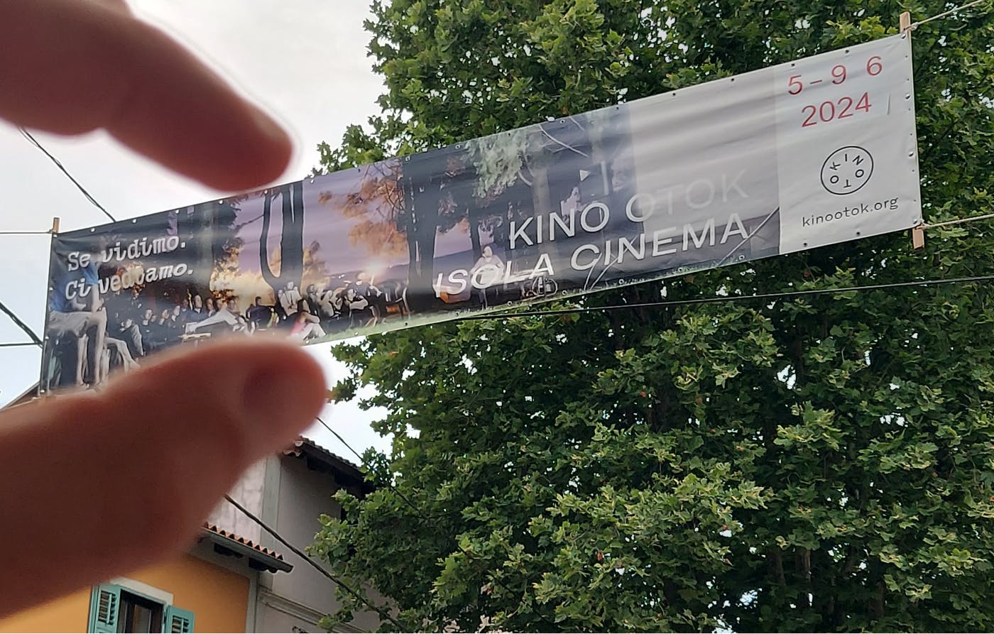 Razpet plakat Kino Otoka 2024 nad ulico, avtor ustvarja iluzijo, kot da ga drži s prsti