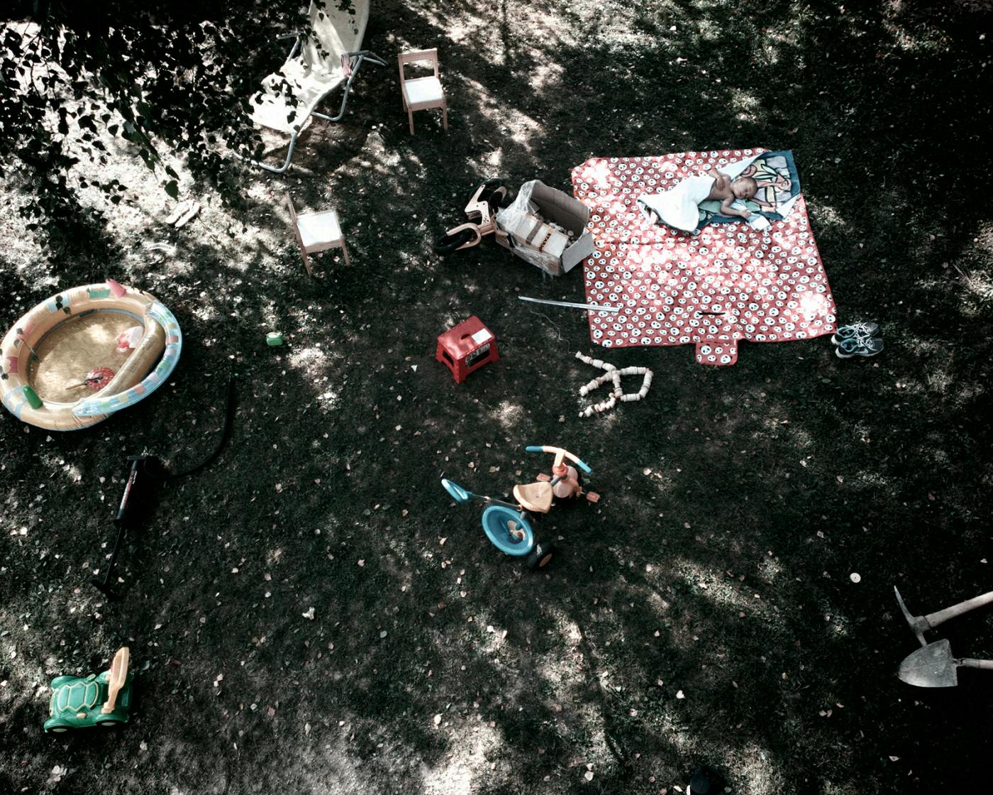 Dvorišče, fotografirano od zgoraj, na njem nekaj otroških igrač in piknik odeja, na kateri spi pokrit otrok