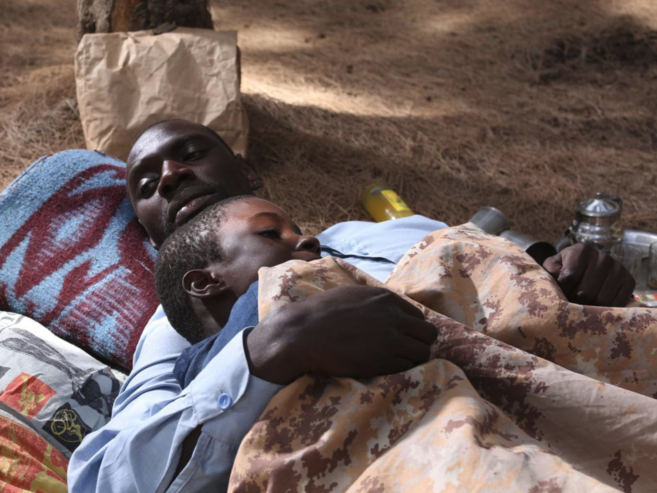 Moški in deček ležita na pesku zavita v barvna ogrinjala in deke. Izsek iz filma Yao.