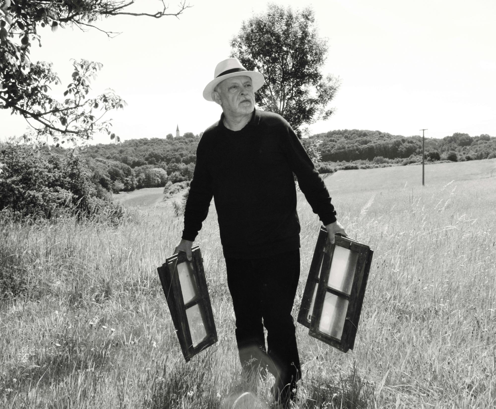 Črno-bela fotografija Ferija Lainška, ki hodi po travniku, v rokah ima okenske okvirje