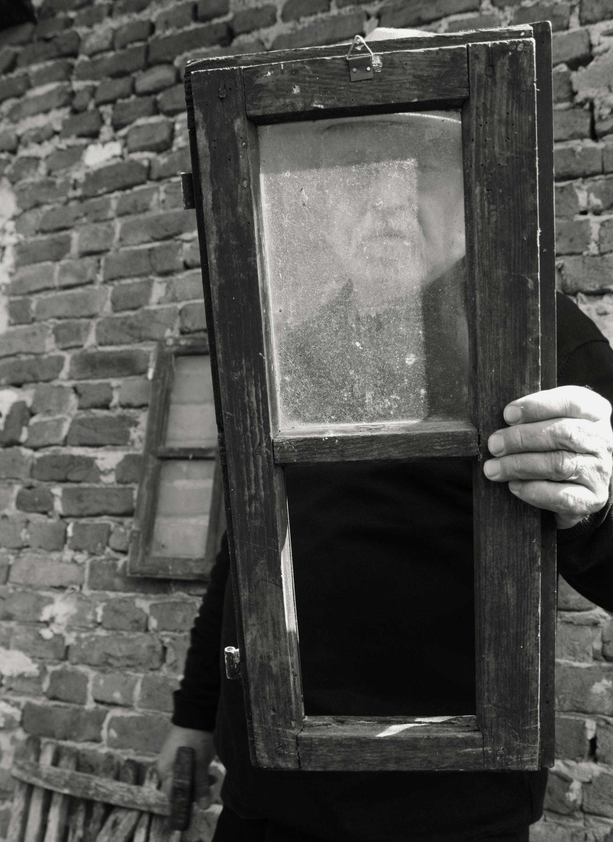 Črno-bela fotografija Ferija Lainščka, ki skozi star okenski okvir gleda v objektiv